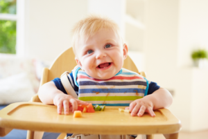 A baby sitting in a high chair with colourful solid foods on the tray in front of them. They are grinning at the camera.