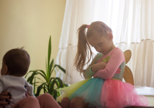 A young girl standing next to her baby brother crosses her arms and pouts towards the camera.